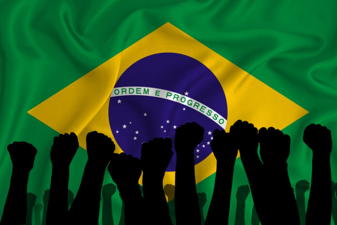 Silhouette of raised arms and clenched fists on the background of the flag of Brazil. The concept of power, power, conflict. With place for your text. Silhouette of raised arms and clenched fists on the background of the flag of Brazil. The concept of power, power, conflict. With place for your text. Brazil military conflict