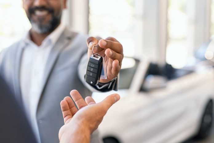 Closeup hand of cardealer giving new car key to customer. Detail of salesman hand giving keys to a client at showroom. Man's hand receiving car keys from african agent in a auto dealership with copy space.