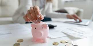 Close up coffee table lot of invoice, financial stats documents, utility bills, household payments, focus on female hand puts coin in piggy bank. Family budget manage, making personal savings concept