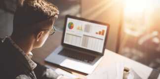 Young finance market analyst in eyeglasses working at sunny office on laptop while sitting at wooden table.Businessman analyze document in his hands.Graphs and diagramm on notebook screen.Blurred