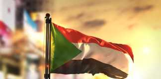 Sudan Flag Against City Blurred Background At Sunrise Backlight