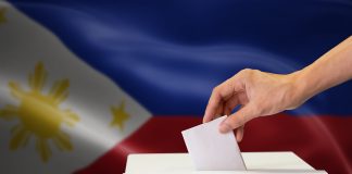 Philippine 2022 Election: Marcos Vs Robredo’s Proxy Campaign Close-up of human hand casting and inserting a vote and choosing and making a decision what he wants in polling box with Philippines flag blended in background