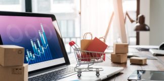 Shopping bags in shopping cart and credit card on laptop with paper boxes on table and sales data economic growth graph on screen, buying and selling services online on network, online shopping and e-commerce concept