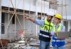 Young attractive construction man and woman in vest with helmet working with laptop, standing on building construction site. Home building project. Engineer foreman discusses with a coworker at workplace.