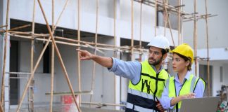 Young attractive construction man and woman in vest with helmet working with laptop, standing on building construction site. Home building project. Engineer foreman discusses with a coworker at workplace.