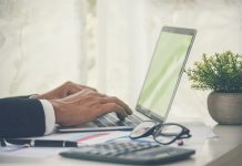 Closeup businessman hands working use laptop finance business document reading report paperwork spreadsheet at office desk. Man hands type keyboard write on office note check financial graph chart