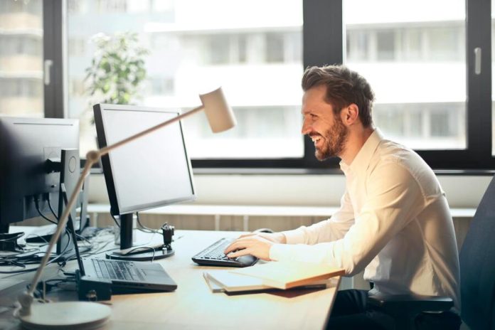 Businessman Sitting on Black Rolling Chair While Facing Black Computer Set and Smiling