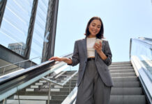 Smiling young Asian business woman standing on escalator using phone