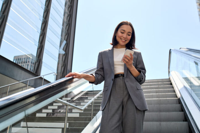 Smiling young Asian business woman standing on escalator using phone