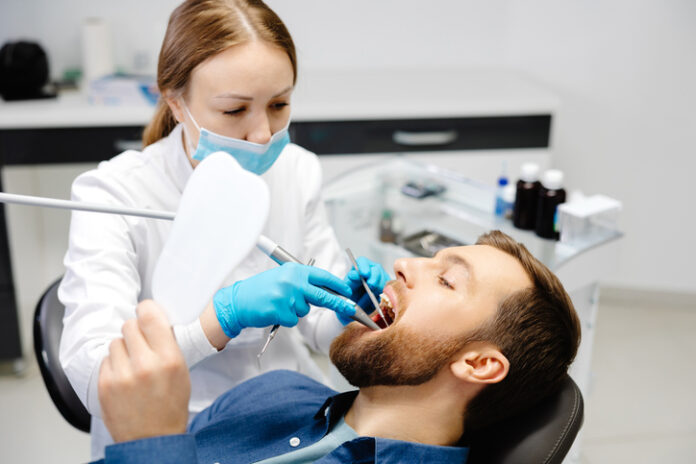 A handsome man is sitting in a chair at a woman's appointment at the dentist's office.