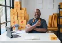 Happy small business owner in apron giving thumbs up in modern office with cardboard boxes, laptop, and notepad.