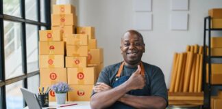 Happy small business owner in apron giving thumbs up in modern office with cardboard boxes, laptop, and notepad.