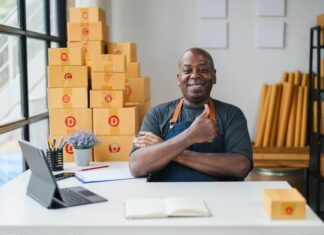 Happy small business owner in apron giving thumbs up in modern office with cardboard boxes, laptop, and notepad.