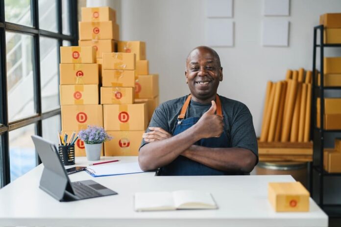 Happy small business owner in apron giving thumbs up in modern office with cardboard boxes, laptop, and notepad.