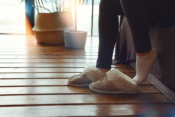 Woman putting on soft plush warm slippers while sitting on sofa at cozy home in cold season