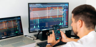 Male trader sits in home office in front of screens with graphs and financial charts, using smartphone, predicts, analyzing and studying the growth or decline of cryptocurrency.