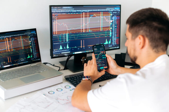 iStock-2148329126 (1) Male trader sits in home office in front of screens with graphs and financial charts, using smartphone, predicts, analyzing and studying the growth or decline of cryptocurrency.