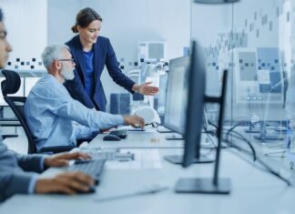 Female Project Supervisor Talks to Male Senior Industrial Engineer who Works on Computer, Talk. In Workshop