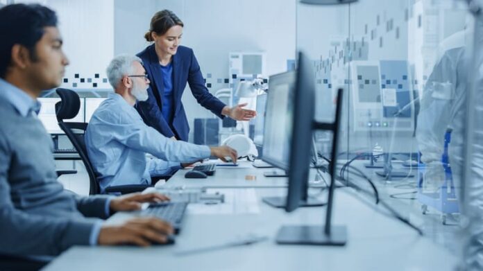Female Project Supervisor Talks to Male Senior Industrial Engineer who Works on Computer, Talk. In Workshop