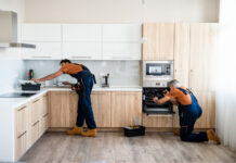 Two handymen, workers in uniform fixing, installing furniture and equipment in the kitchen, using screwdriver indoors.