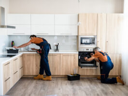 How to Save Big on Renovations with Wholesale Cabinets Two handymen, workers in uniform fixing, installing furniture and equipment in the kitchen, using screwdriver indoors.