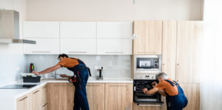 Two handymen, workers in uniform fixing, installing furniture and equipment in the kitchen, using screwdriver indoors.