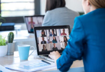 Back view of two business women speaking on video call with diverse colleagues on online briefing with laptop in the office.