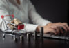 Close up shot of woman typing on laptop with mini shopping cart and coins stack.