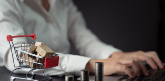 Close up shot of woman typing on laptop with mini shopping cart and coins stack.