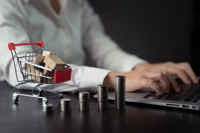 Close up shot of woman typing on laptop with mini shopping cart and coins stack.