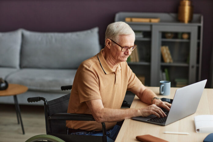 Senior man typing on laptop Senior man with disability sitting on wheelchair and typing on laptop