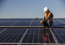 A supervisor is crouching on the rooftop surrounded by solar panels and running tests on the laptop.