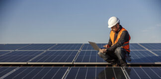 A supervisor is crouching on the rooftop surrounded by solar panels and running tests on the laptop.