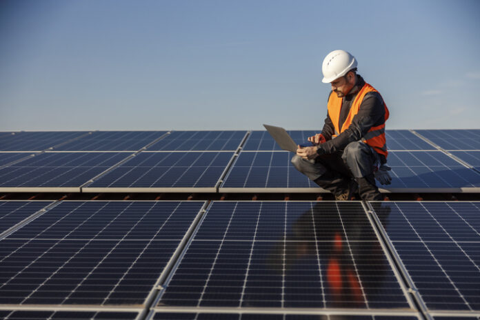 A supervisor is crouching on the rooftop surrounded by solar panels and running tests on the laptop.