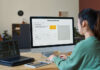 Young female designer sitting in front of desktop computer with AI image generator on screen and typing on keypad