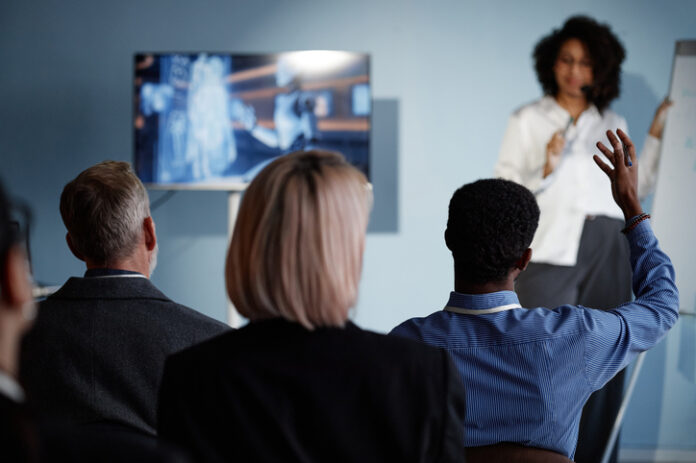 Back view of Black male business meeting attendee raising hand to ask question to colleague during presentation on latest updates in AI technologies