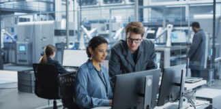 Female Engineer Collaborating on a Project, Using Computer and Talking in a Factory with Robotic Arms Producing Modern High Tech Devices.