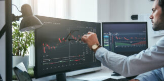 Young trader is pointing at graphs on computer screen and analyzing data while working in his modern office.