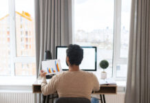 Back view of the serious concentrated determined young bearded office worker in business casual clothes, sitting at the desk, typing on laptop.