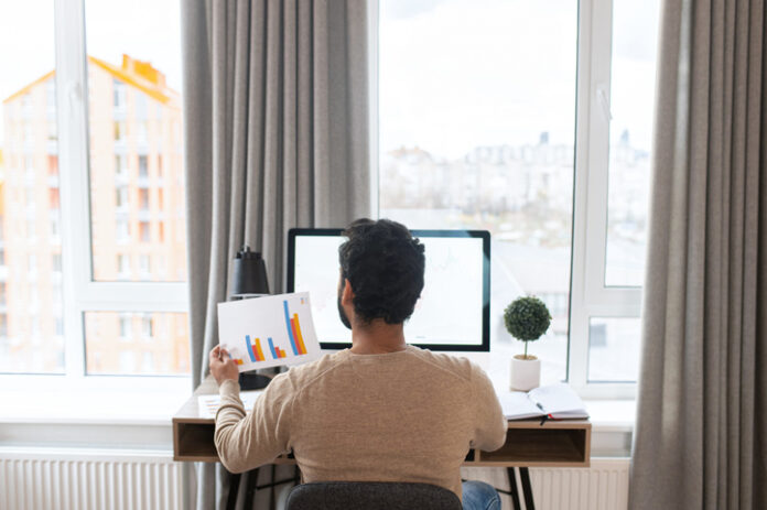 Back view of the serious concentrated determined young bearded office worker in business casual clothes, sitting at the desk, typing on laptop. Businessman professional is working on project Back view of the serious concentrated determined young bearded office worker in business casual clothes, sitting at the desk, typing on laptop.