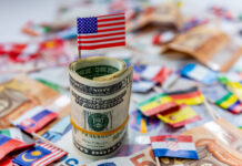 Reconfiguring US Hegemony: Militarism, Empire, and the Crisis of Capitalist Accumulation A roll of US dollars with the American flag on top of a other currencies and country flags.
