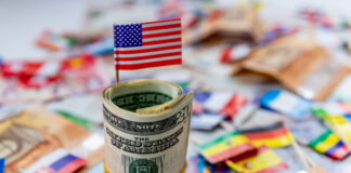 A roll of US dollars with the American flag on top of a other currencies and country flags.