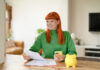 Young woman with red hair managing finances while using her smartphone and smiling in a cozy living room