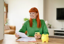 Young woman with red hair managing finances while using her smartphone and smiling in a cozy living room