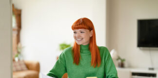 Young woman with red hair managing finances while using her smartphone and smiling in a cozy living room