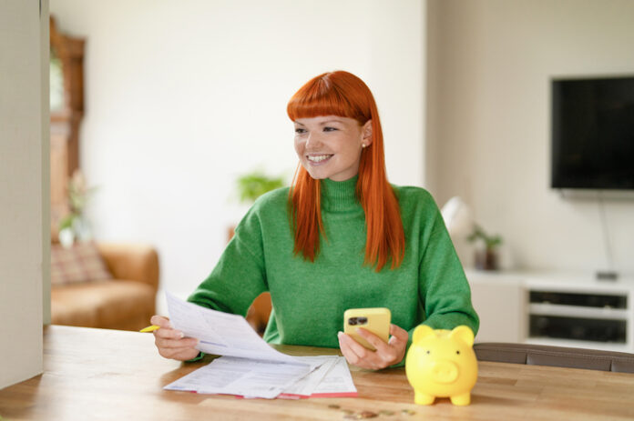 Young woman with red hair managing finances while using her smartphone and smiling in a cozy living room