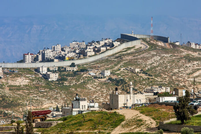 iStock-482351723 (1) Small village and palestinian town on the hill behind israeli separation barrier on the West Bank in Israel.