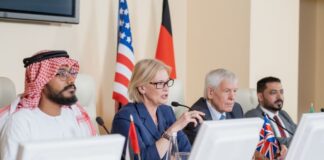 Blond mature female delegate making speech for audience while sitting by table between several foreign colleagues