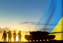 Silhouettes of a soldiers and a main battle tank on a battlefield with Ukraine flag against the sunset.