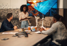 Portrait of multicultural ecology engineer standing with solar panels model in hands at boardroom and having seminar about clean and renewable energy development in front of diverse team of experts.